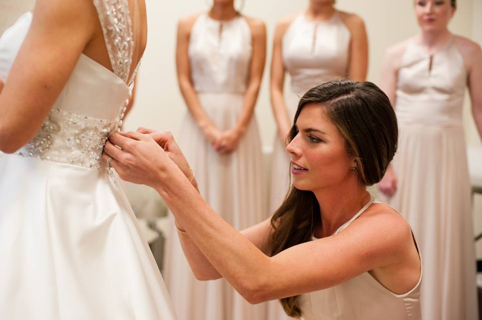 A maid of honor adjusts a bride's dress while her bridesmaids look on.