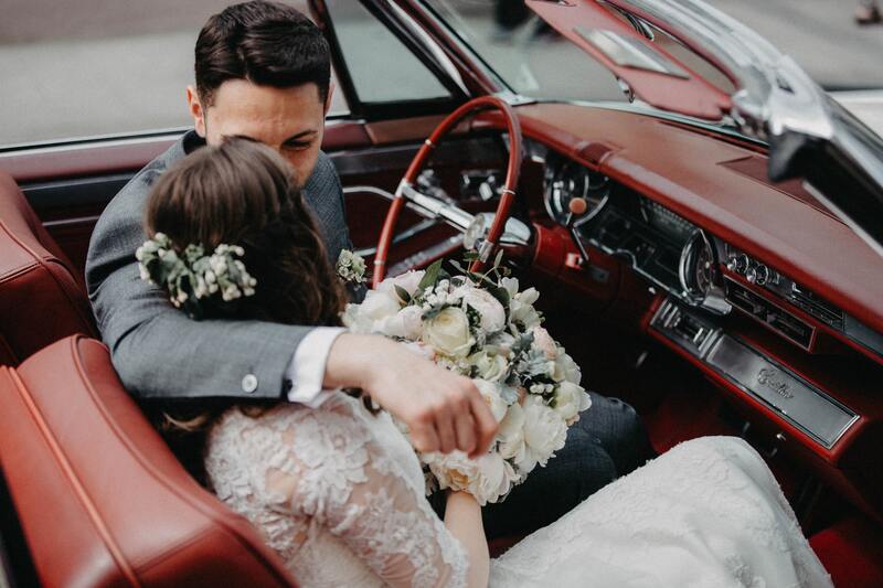 A bride and groom share a kiss in the front seat of a classic car.