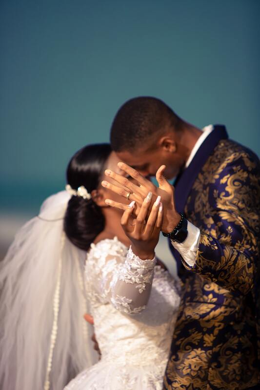 A bride in a wedding dress kisses a groom in a purple and gold jacket while they hold their hands up to the camera, showing off their wedding bands